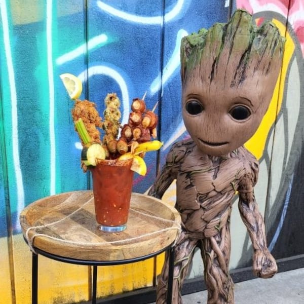 A Groot statue stands next to a Bloody Mary garnished with fried seafood and bacon-wrapped items.
