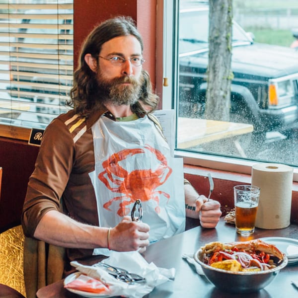 A man with a beard and glasses wearing a crab bib, holding seafood crackers, with a bowl of seafood boil.