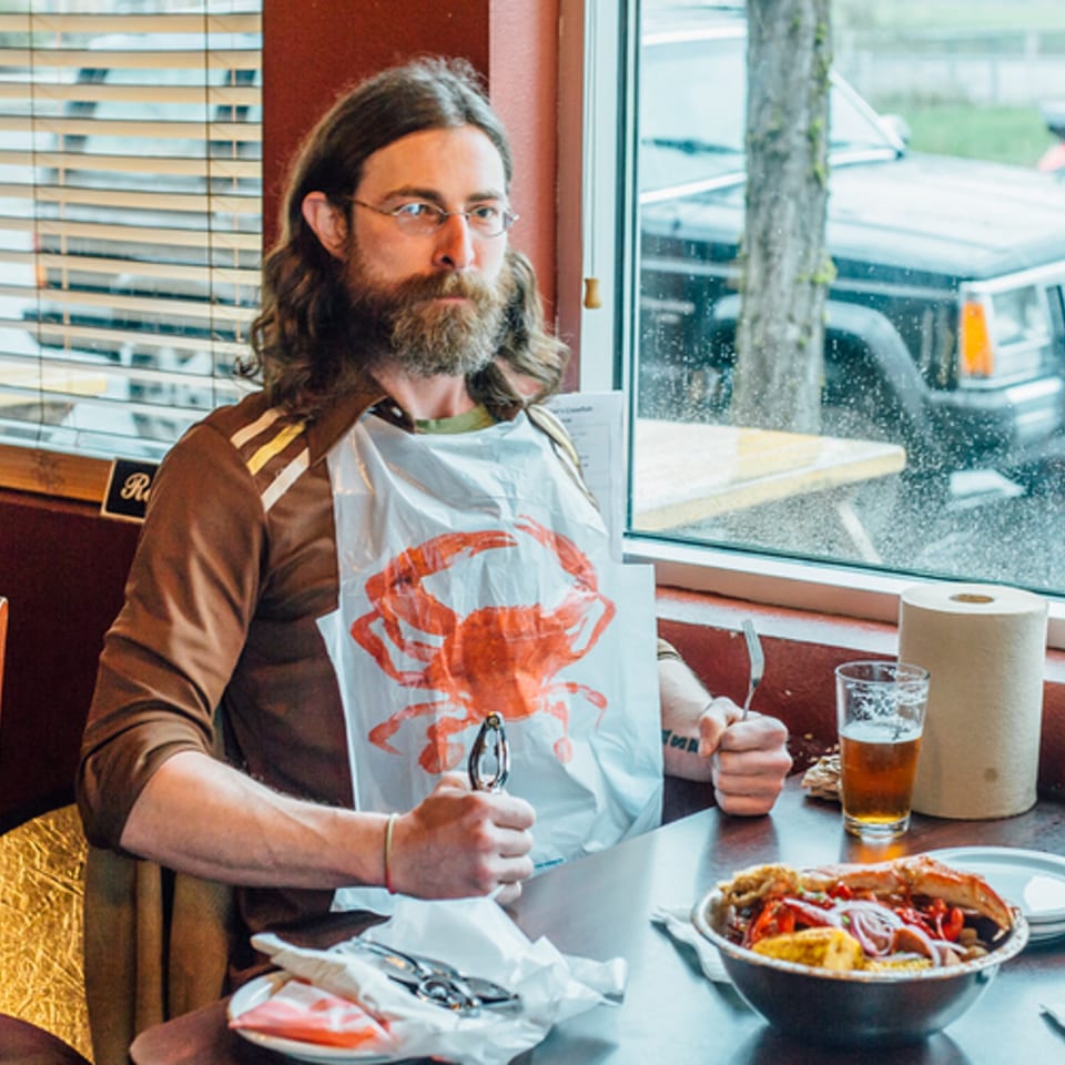 A man with a beard and glasses wearing a crab bib, holding seafood crackers, with a bowl of seafood boil.