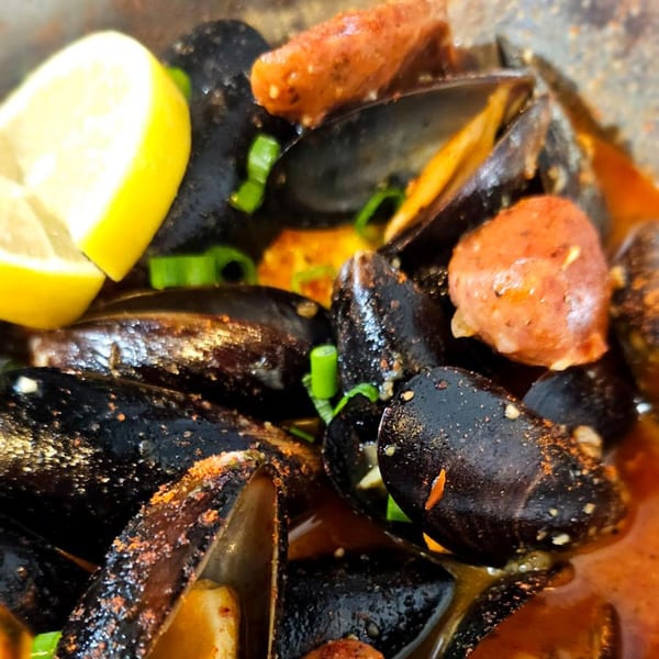 Steamed mussels with sliced sausage, green onions, and a lemon wedge in a spicy broth.