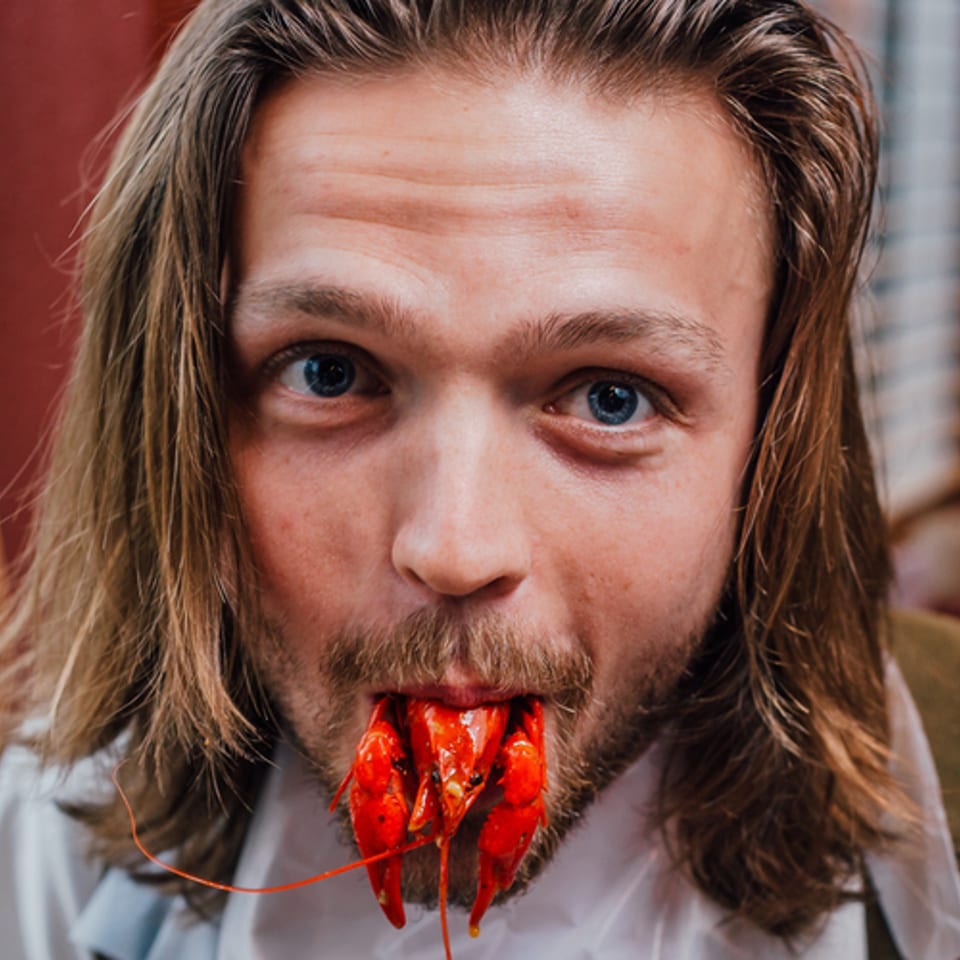 Man with long hair and beard, wearing a bib, holding a crawfish in his mouth.