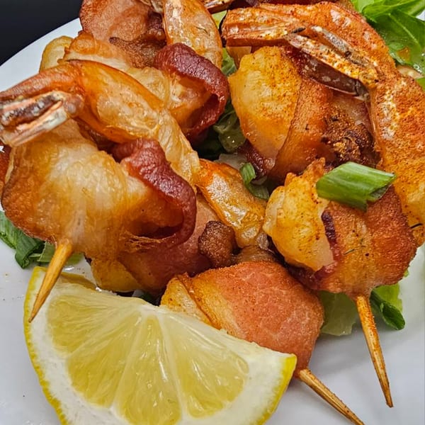 Bacon-wrapped shrimp skewers with a lemon wedge and green onions on a white plate.